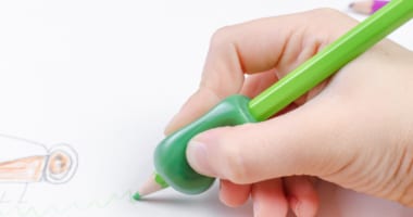A child's hand drawing with a green pencil using an adapted handle for wider grip.