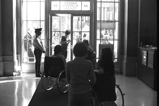 in the foreground a woman in a wheelchair has her back to us along with other members of the sit-in. In front of her is a large glass door with a security guard, outside are additional protestors