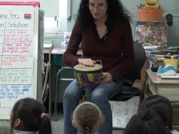 Teaching Artist with drum sitting in front of students. One hand is tapping on the drum.