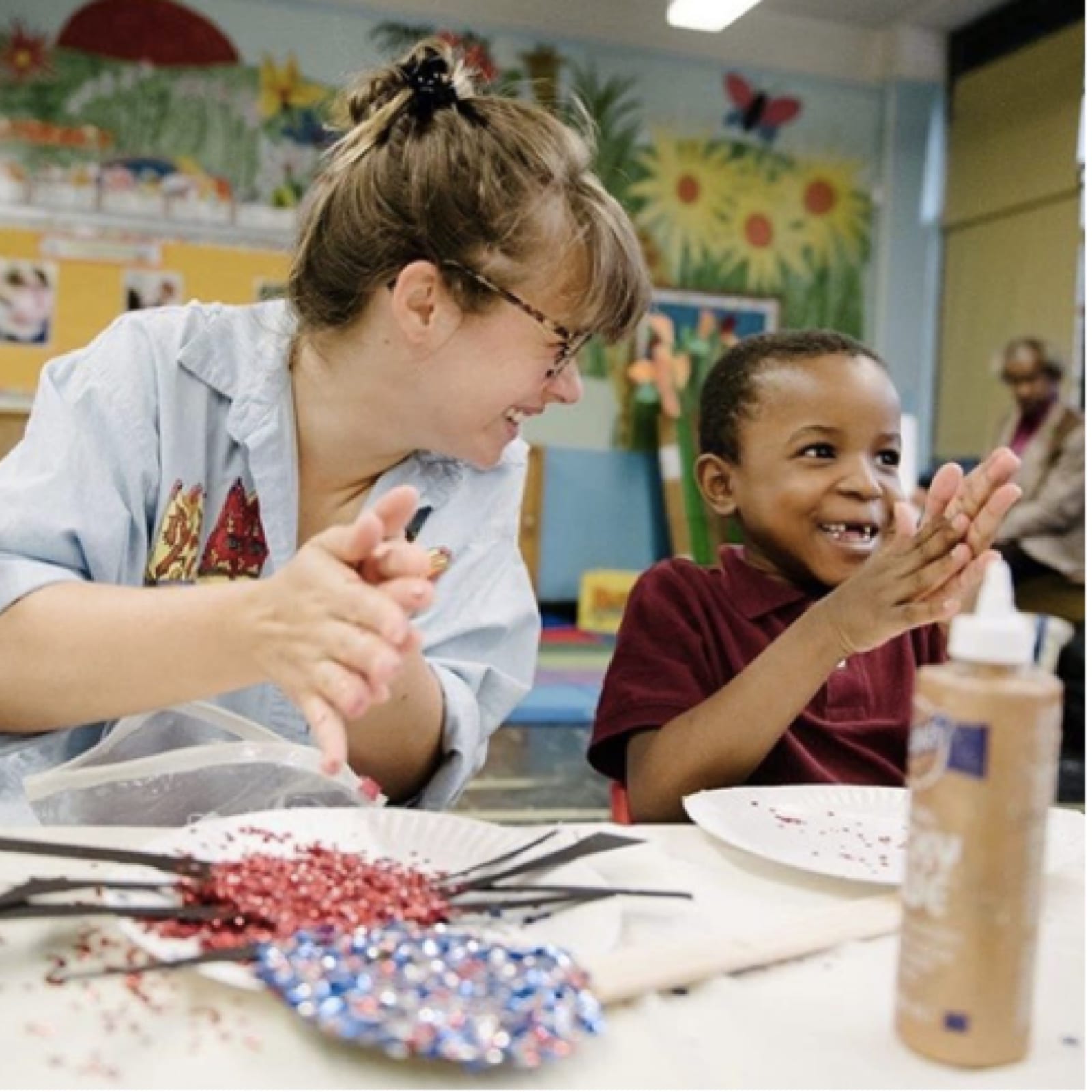 Teaching artist and young student sit at a table and rub glitter between their hands, smiling.