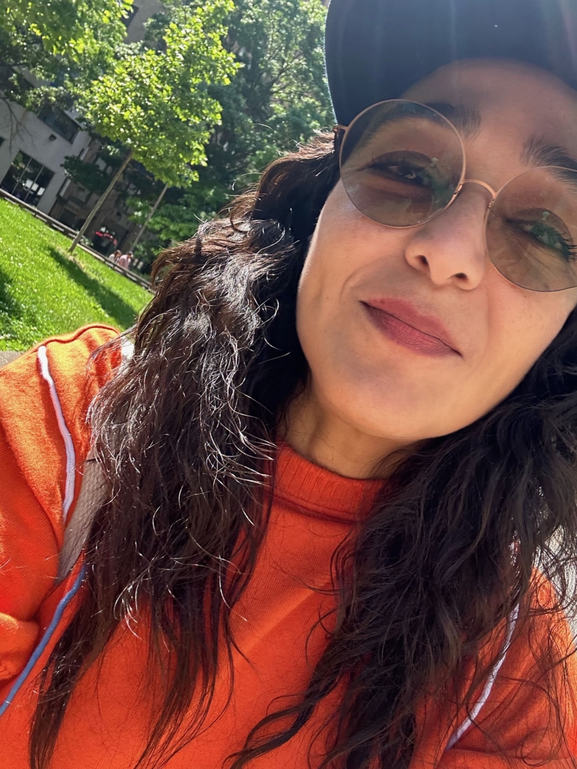 A photo of a smiling Odalis looking at the camera wearing circular sunglasses and a red top. A green lawn is in the background.