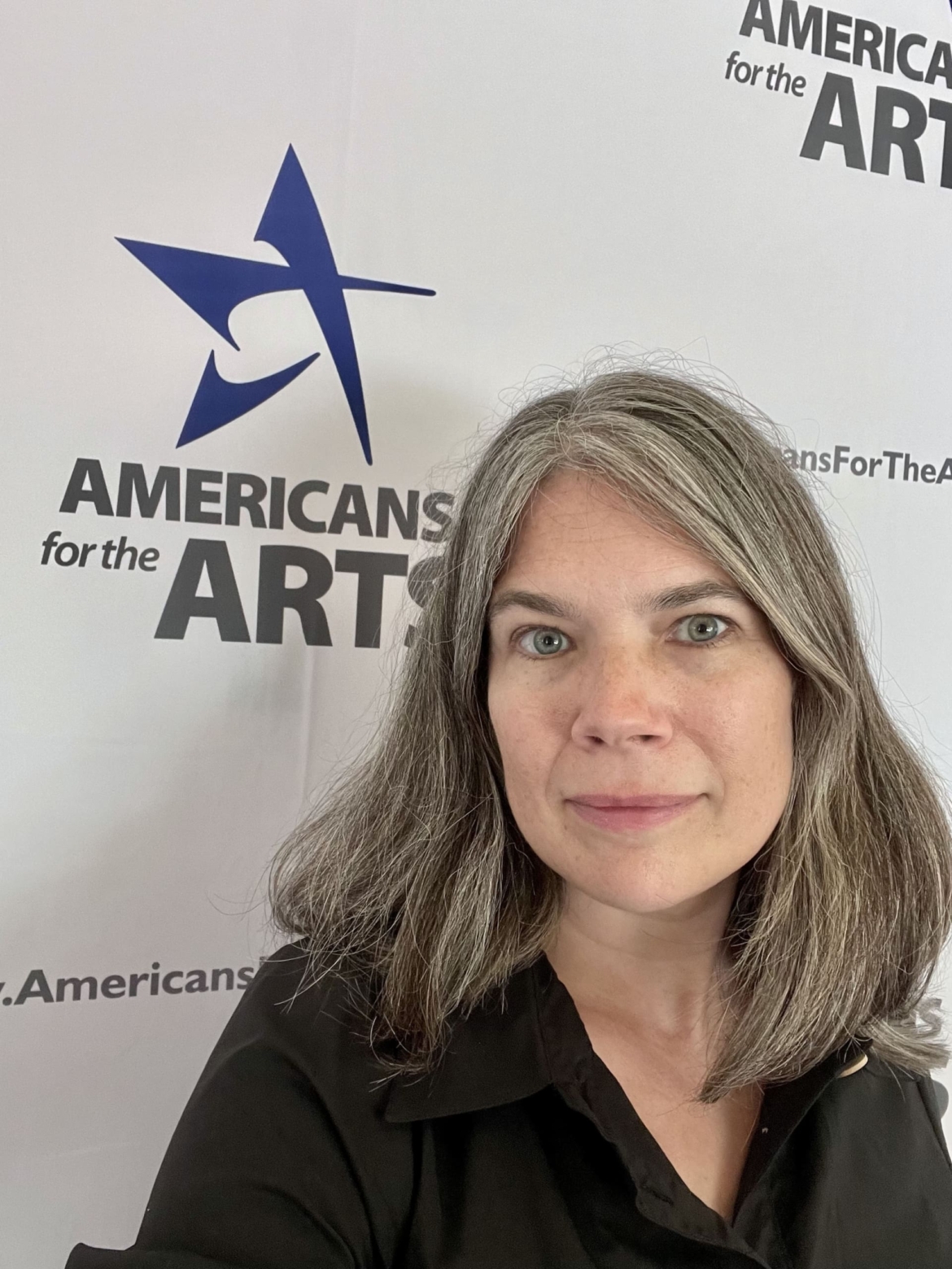 A photo of Erin wearing a back button up shirt, looking directly ahead, standing in front of an American for the Arts banner.